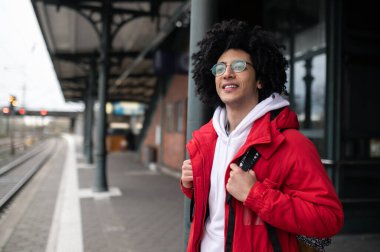 Treni bekliyorum. Kırmızı ceketli ve sırt çantalı bir adam platformda treni bekliyor.