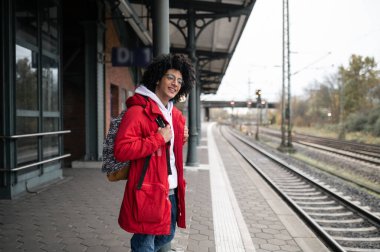 Treni bekliyorum. Kırmızı ceketli ve sırt çantalı bir adam platformda treni bekliyor.