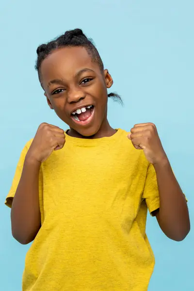 非常快乐的非洲裔美国小男孩与紧紧抓住的拳头在蓝色背景下欢欢喜喜 免版税图库照片