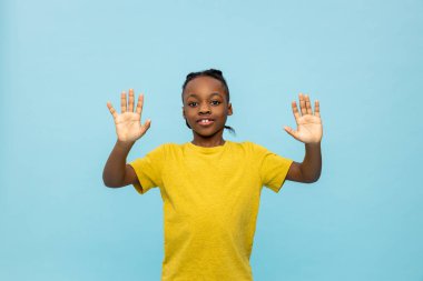 Stüdyoda bir çocuk. Sarı tişörtlü çocuk mavi arka planda duruyor ve el kol hareketi yapıyor.