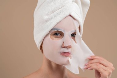 Yüzünde gençleştirici maskesi olan genç bir kadın kahverengi arka planda izole edilmiş.