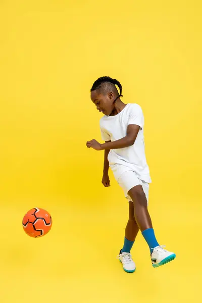 黄色の背景に隔離されたフライングキックをしているサッカーボールを持つ若いアフリカ系アメリカ人の少年 ロイヤリティフリーのストック写真