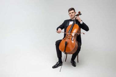 Tutkulu çellocu, beyaz arka planda izole edilmiş bir şekilde koltukta oturmuş konser veriyor.