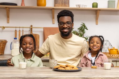 Happy African father and daughter eating cookies and drinking milk at home kitchen clipart
