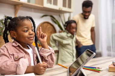 Afro-Amerikalı çocuk mutfak masasında oturmuş dijital tabletinin yardımıyla ödevini tamamlıyor.