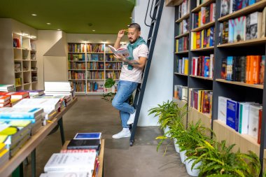 Kitap okuyordum. Genç Asyalı adam kitap raflarının yanında duruyor ve kitap okuyor.