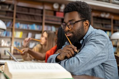 Kütüphanede. Kıvırcık saçlı Afrikalı Amerikalı adam kütüphanede düşünceli görünüyor.
