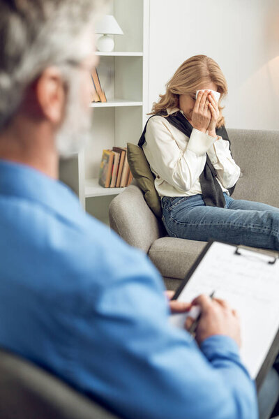 Unhappy woman sitting on couch in counseling office and complaining about his troubles to male psychologist while he taking notes on clipboard