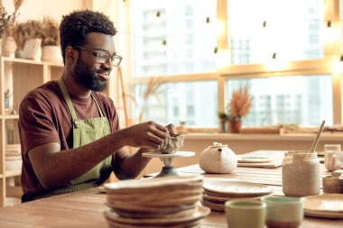 Çömlek. Kıvırcık saçlı adam masada oturuyor, kille çalışıyor ve ilgili görünüyor.
