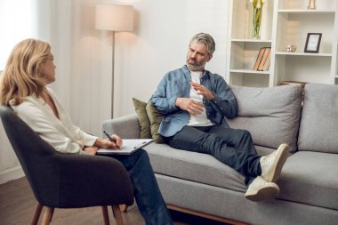 Mutsuz bir adam danışmanlık ofisinde kanepede oturup kadın psikoloğa dertlerinden yakınırken o da not alıyor.