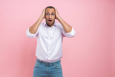 Şok olmuş beyaz gömlekli adam ellerini pembe arka planda kapalı tutuyor.