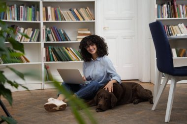 Arkadaşınla çalış. Dizüstü bilgisayarı olan kıvırcık saçlı bir kadın yerde oturuyor, yanında da bir köpek.