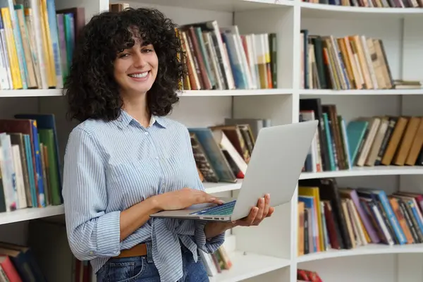 Kütüphanedeki kadın. Kütüphanedeki kitap raflarının yanında dizüstü bilgisayarı olan beyaz bir kadın.