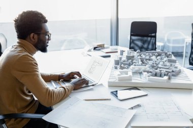 İşyerinde teknik taslak ve çizimler kullanarak dizüstü bilgisayar üzerinde çalışan siyah bir mimar.