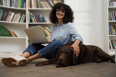 Kıvırcık saçlı kız öğrenci üniversite kütüphanesinde köpekle çalışırken dizüstü bilgisayar kullanıyor.