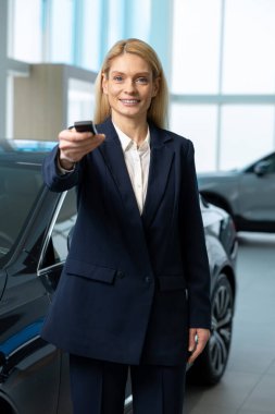 Satış. Zarif takım elbiseli sarışın kadın arabanın yanında elinde anahtarla duruyor.