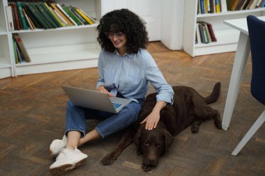 Kıvırcık saçlı kız öğrenci üniversite kütüphanesinde köpekle çalışırken dizüstü bilgisayar kullanıyor.