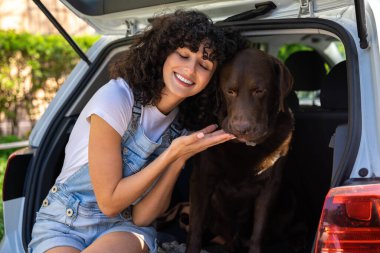 Arkadaşız. Genç kıvırcık saçlı kadın arabanın bagajında otururken köpeğine sarılıyor.