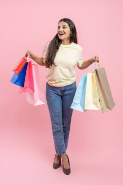 Normal tişört ve kot pantolon giyen bir kadının uzun bir portresi pembe arka planda izole edilmiş alışveriş çantaları taşıyor.
