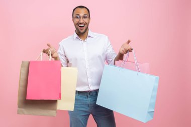 Pembe arka planda izole edilmiş alışveriş torbaları alırken gömlek ve gözlük takan aşırı neşeli Afrikalı Amerikalı bir adam.