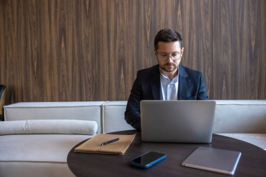 Sakallı başarılı girişimci resmi bir iş süitinde koltukta oturuyor ve iç dizüstü bilgisayarla oturuyor.