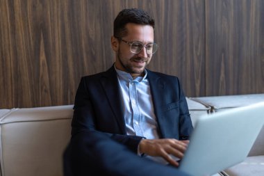 Sakallı başarılı girişimci resmi bir iş süitinde koltukta oturuyor ve iç dizüstü bilgisayarla oturuyor.