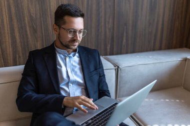 Sakallı başarılı girişimci resmi bir iş süitinde koltukta oturuyor ve iç dizüstü bilgisayarla oturuyor.