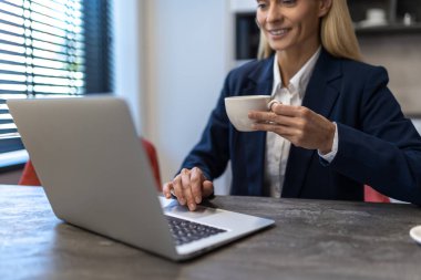 Güzel beyaz iş kadını dizüstü bilgisayarda çalışıyor ve iş yerinde sıcak kahvenin tadını çıkarıyor.