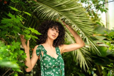 Koyu kıvırcık saçlı kadın tropikal yaprakların arka planında yaz tazeliğini ifade eder.