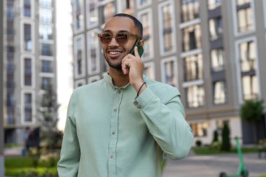 Açık yeşil tişörtlü Afro-Amerikalı açık havada akıllı telefondan konuşuyor.