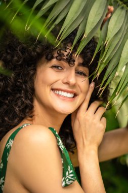Kıvırcık saçlı, gülümseyen kadın model. Arka planda taze egzotik yapraklar var.