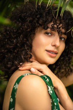 Yeşil yaprakların arasında siyah kıvırcık saçlı güzel bir kadın model şık bir elbise içinde mutlu görünüyor.