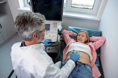 İşteki doktor. Erkek, gri saçlı profesyonel. Ultrasonografi cihazı üzerinde çalışıyor ve kendinden emin görünüyor.