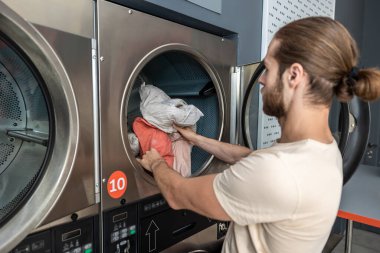 Çamaşırhanede çamaşır makinesini yükleyen ev işleriyle meşgul genç bir adam.