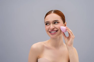 Yüz bakımı. Güzel, kızıl saçlı, beyaz bir kadın yüz süngeri kullanıyor ve gülümsüyor.