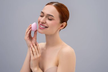 Yüz bakımı. Güzel, kızıl saçlı, beyaz bir kadın yüz süngeri kullanıyor ve gülümsüyor.