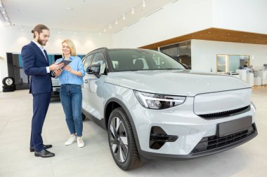 Sarışın kadın otomobil dükkanındaki bayiyle araba alışverişi yapıyor.