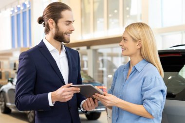 Mavi gömlekli kadın oto galerisindeki müşteri hizmetleri müdürüyle lüks araç satın alma işini sonuçlandırıyor.