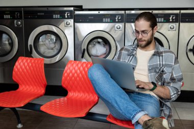Çamaşırhanede çamaşır makinesini beklerken kablosuz internet ve dizüstü bilgisayar kullanan beyaz bir adam.