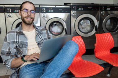 Beyaz tenli yakışıklı, çamaşırhanede laptoptan internete giriyor.