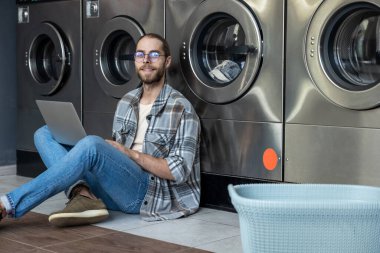 Kafkasyalı bir adam laptopla birden fazla iş yapıyor ve ev işleriyle uğraşıyor yerde oturmuş çamaşırhanede bekliyor.