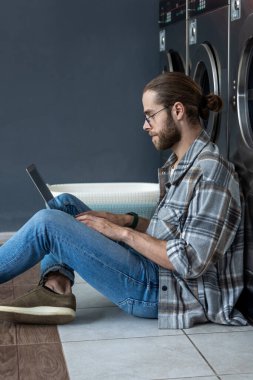 Çamaşırhanede çamaşır makinesini beklerken kablosuz internet ve dizüstü bilgisayar kullanan beyaz bir adam.