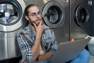 Kamu çamaşırhanesinde çamaşır beklerken dizüstü bilgisayarda çalışan dalgın beyaz adam.