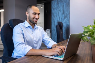 Mavi tişörtlü sakallı adam ofiste dizüstü bilgisayarla çalışıyor.