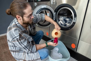 Beyaz sakallı bir adam çamaşır makinesini ve deterjanı kendi kendini yıkamada kullanıyor.