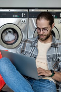 Beyaz adam, halka açık çamaşırhanede beklerken elinde defterle online alışveriş yapıyor.