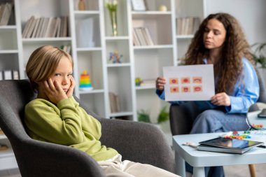 Konuşma terapisi. Uzun saçlı, genç bir konuşma terapisti sarışın bir çocukla çalışıyor ve ikisi de işin içinde gibi görünüyor.