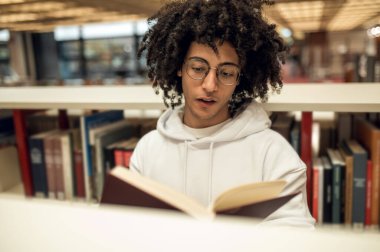 Gülümseyen neşeli üniversite öğrencisi kütüphanede kitap okuyor.