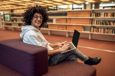 Kıvırcık saçlı erkek öğrenci, kütüphane kampüsünde dizüstü bilgisayarla diploma projesi üzerinde çalışıyor.