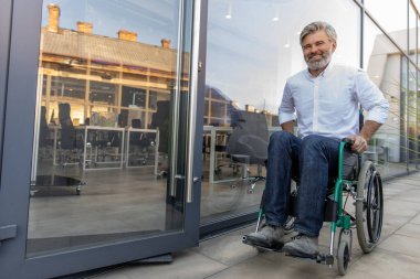 Engelli. Ofis bölgesinde tekerlekli sandalyede oturan gri saçlı, olgun bir adam.